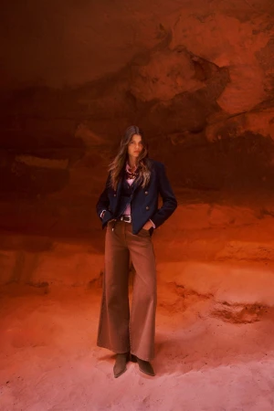 Model Mathilde Henning in a navy jacket and wide-leg brown trousers standing inside a red sandstone cave.