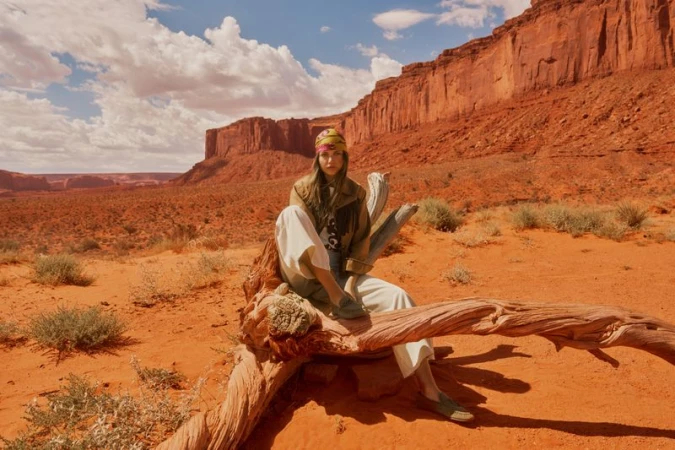 Model Mathilde Henning sitting on driftwood in a red rock desert landscape wearing white wide-leg trousers and a headscarf.