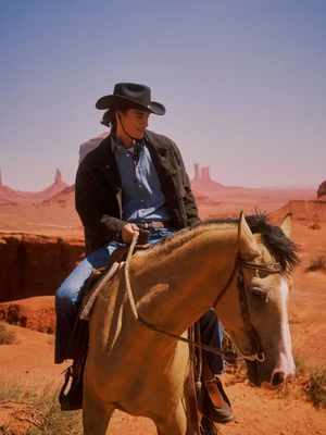 James Turlington wearing a black cowboy hat and denim shirt riding a horse across a desert landscape.