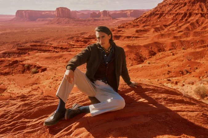 Model James Turlington seated on red desert rock wearing a brown jacket, beige trousers, and black leather shoes.