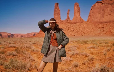 Model Mathilde Henning shielding her eyes in a desert landscape wearing an olive quilted coat and patterned skirt.