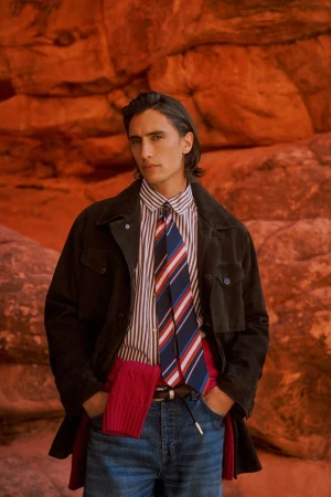Model James Turlington wearing a dark jacket, striped shirt, tie, and blue jeans against a red rock backdrop.