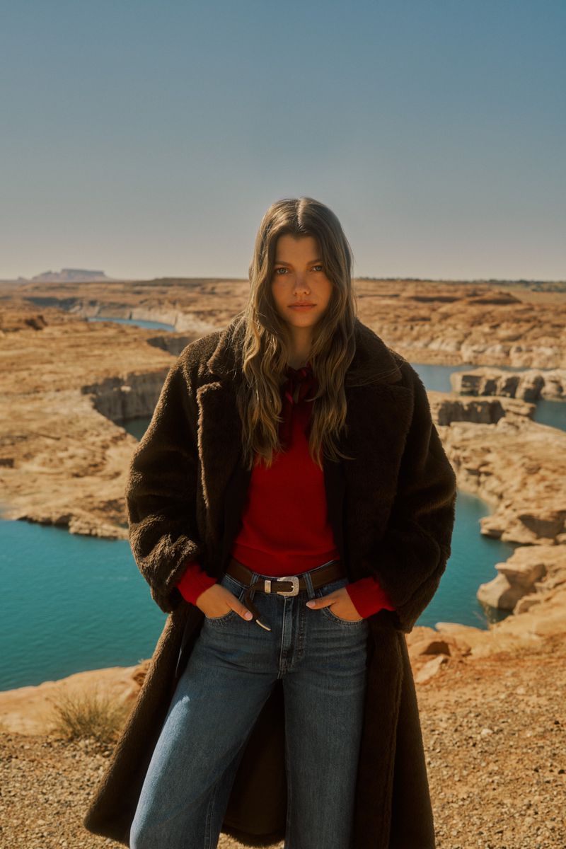 Model Mathilde Henning posing in a dark brown teddy coat, red sweater, and blue jeans against a desert canyon and blue lake.