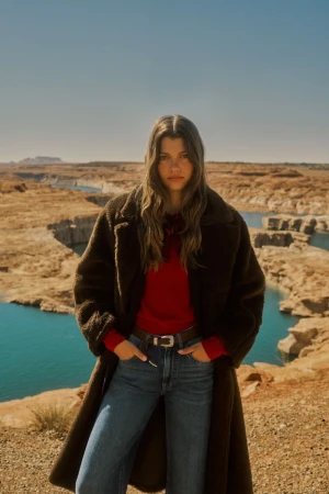 Model Mathilde Henning posing in a dark brown teddy coat, red sweater, and blue jeans against a desert canyon and blue lake.