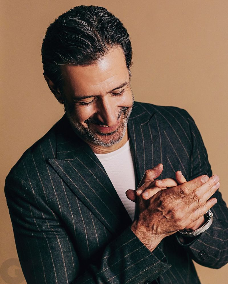 Portrait of a middle-aged man in a pinstripe suit smiling and clasping his hands together indoors in front of a tan background.