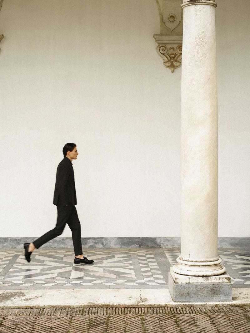 Model wearing a tailored black suit and loafers walking past a marble column in a palace courtyard.