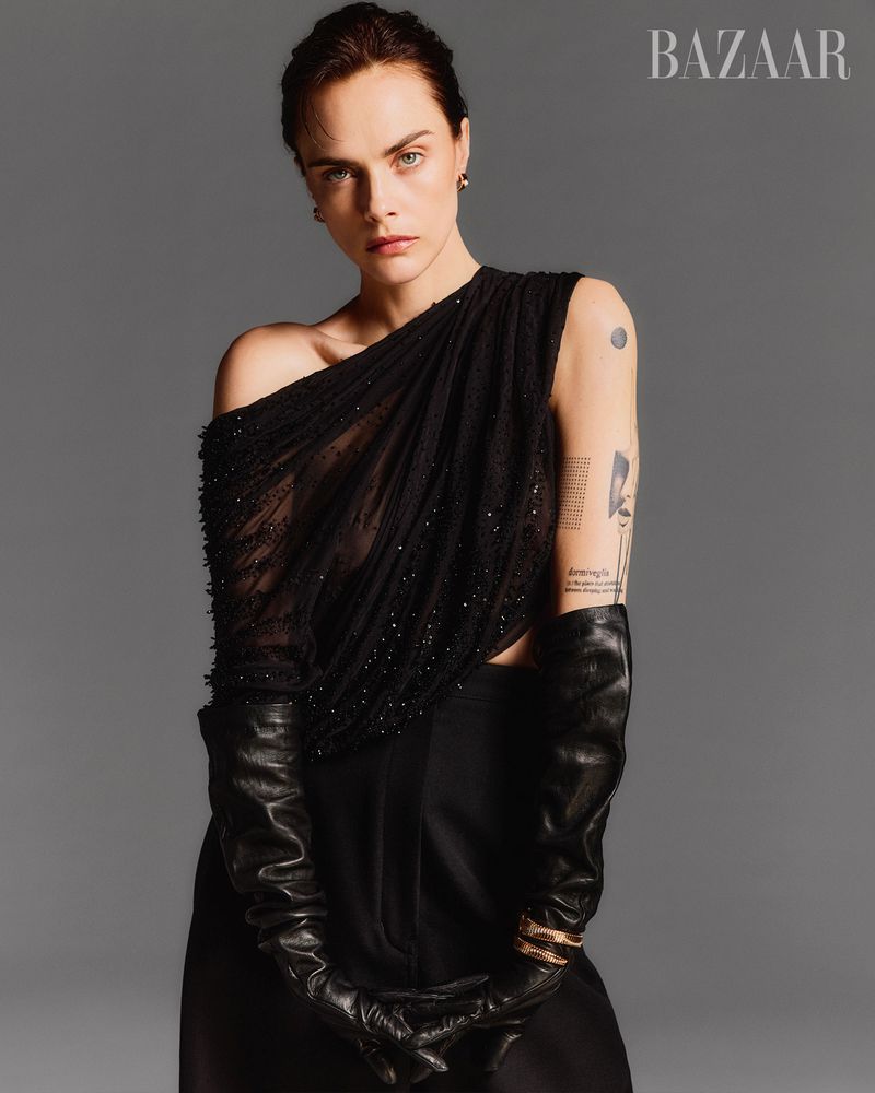 Model Cara Delevingne wearing a black beaded one-shoulder top and long leather opera gloves against a plain grey studio background.