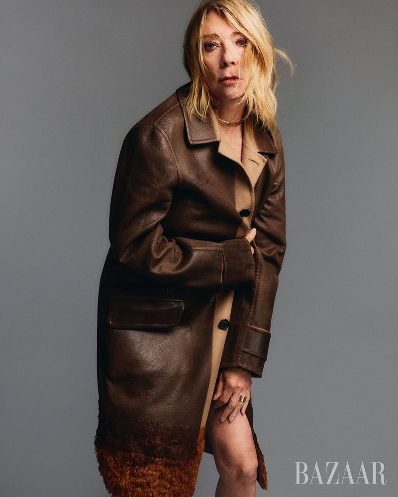 Kim Gordon posing in a long brown leather coat with fur trim against a neutral grey studio backdrop.