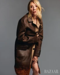 Kim Gordon posing in a long brown leather coat with fur trim against a neutral grey studio backdrop.