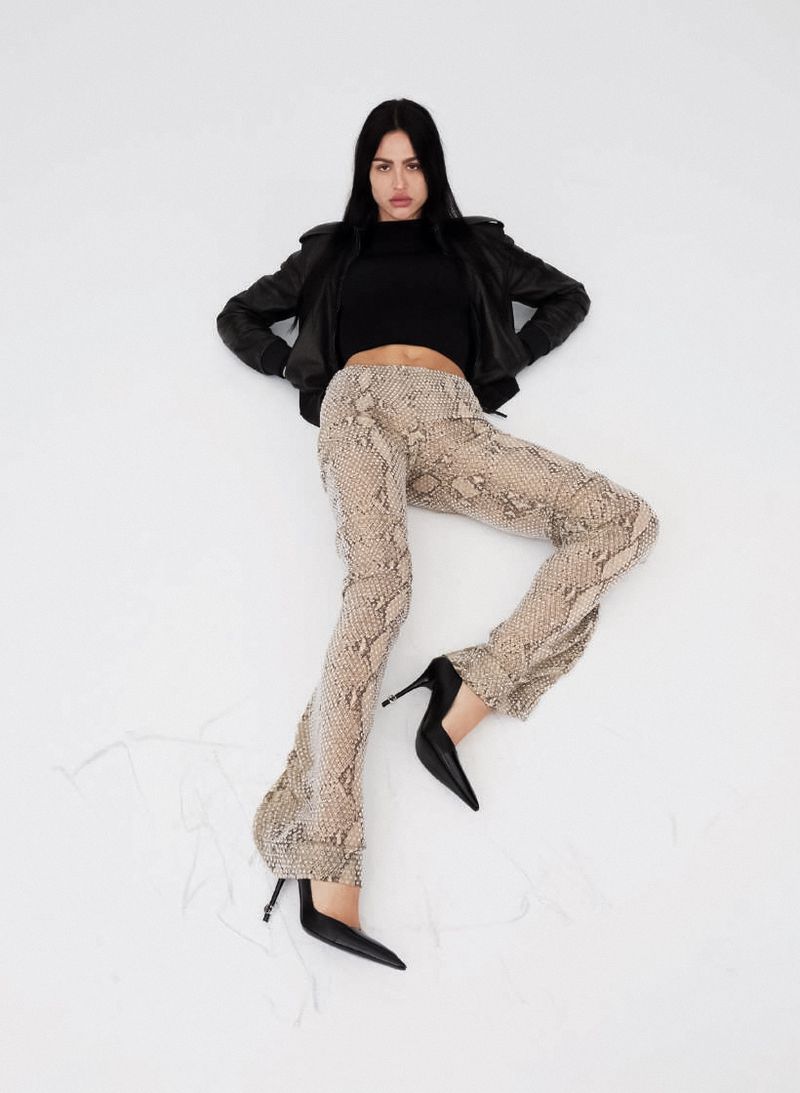 Amelia Gray lies on a white seamless studio background wearing snakeskin print trousers, a black cropped top, and black high heels.