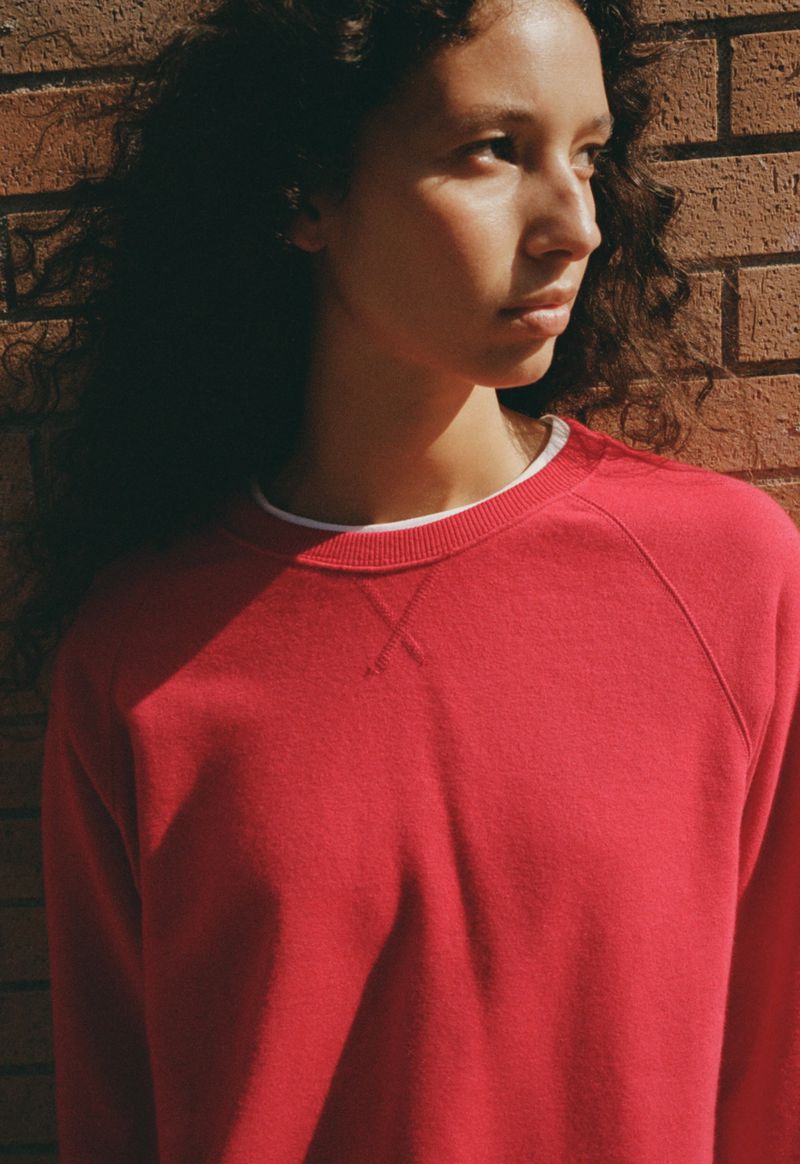 Model Jenn du Puy in a red crewneck sweatshirt standing in direct sunlight against an outdoor brick wall.