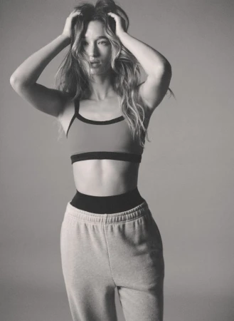 Model Jenn du Puy posing with hands in her hair wearing a sports bra and grey sweatpants against a studio backdrop.