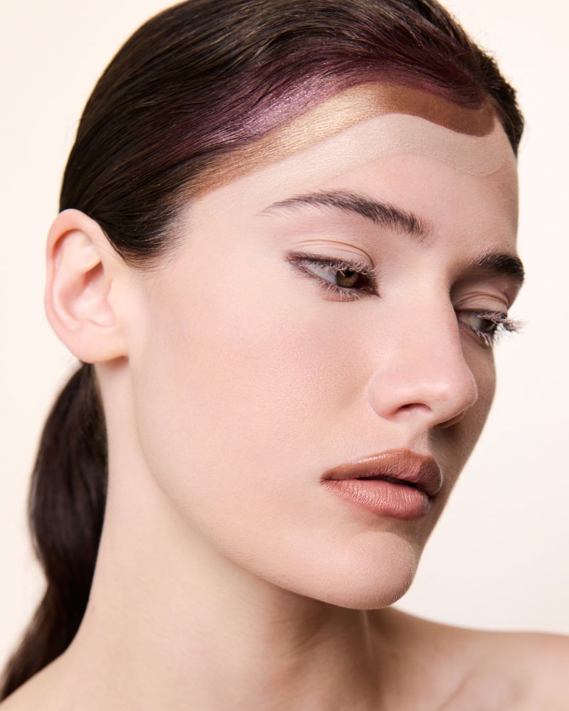 Close-up beauty portrait of a woman with graphic metallic and nude paint waves across her forehead against a pale background.