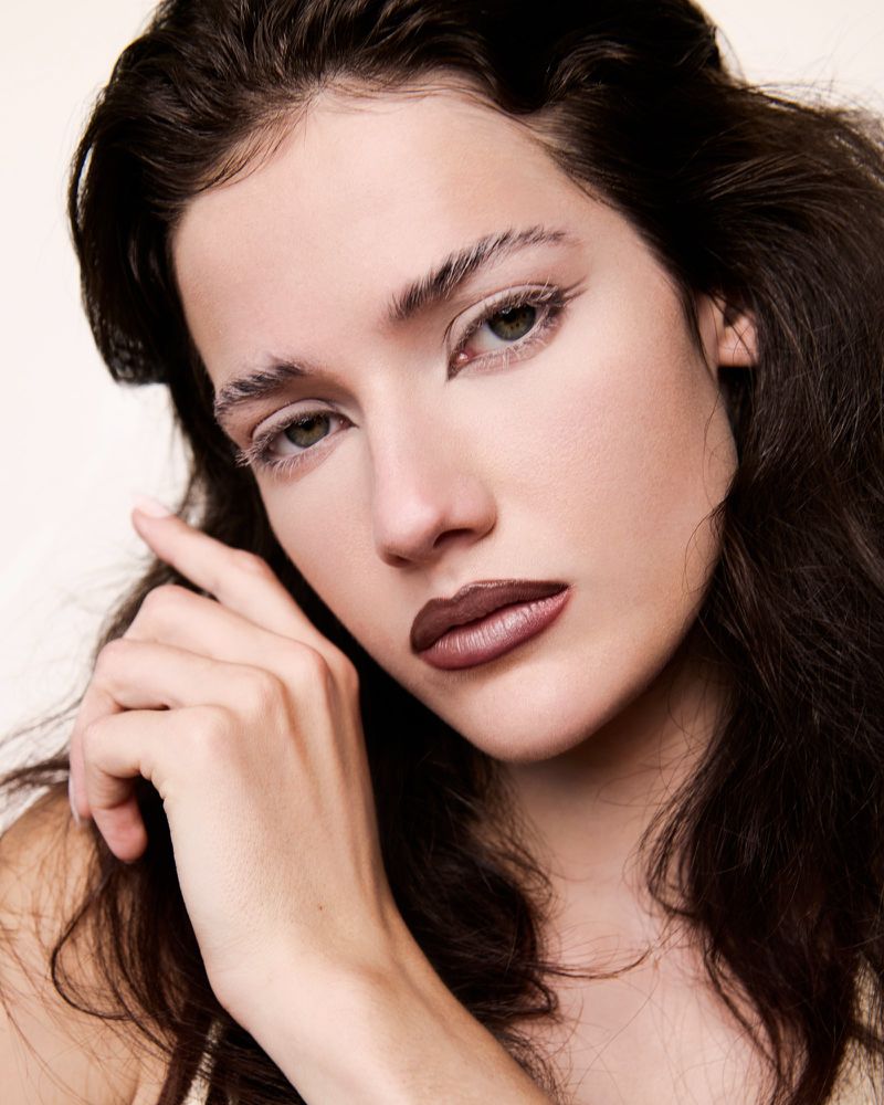 Close-up beauty portrait of a woman with dark wavy hair, white graphic eyeliner, and dark matte lipstick resting her hand near her face.