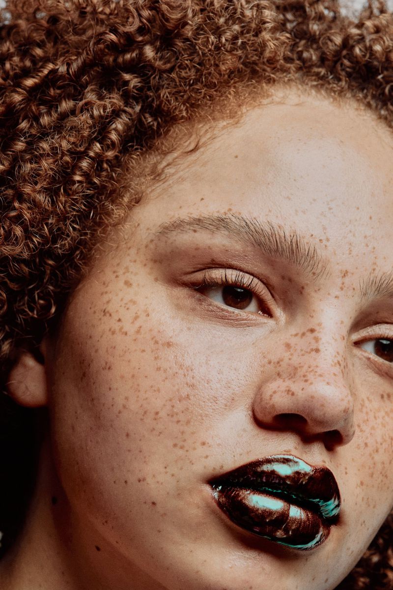 Close-up beauty portrait of a woman with freckles, curly auburn hair, and high-gloss black lipstick with metallic teal sheen.