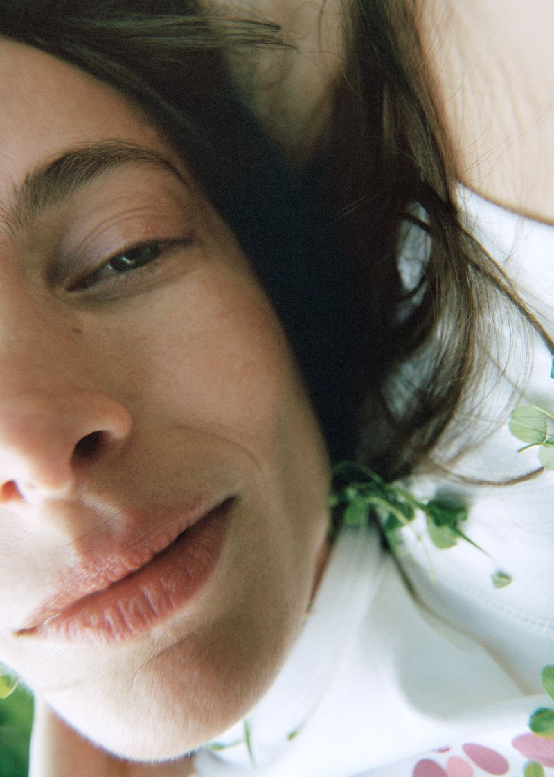 Close-up portrait of an adult woman with brown hair, fair skin, and natural makeup, accented with green clover sprigs and soft lighting.