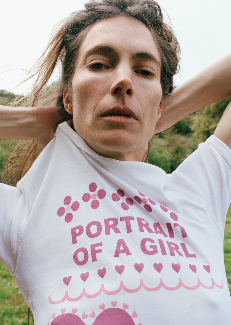 Portrait of an adult woman with arms raised behind her head, wearing a graphic tee with flowers, hearts, and text in an outdoor setting.