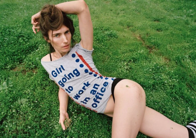 Model Jessica Miller lying on green grass wearing a grey graphic t-shirt with slogan text and black briefs.