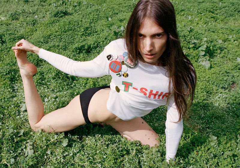 Woman in T-shirt lies on lawn. She has brown hair and wears a white long-sleeved T-shirt with colorful badges. Black shorts are visible.