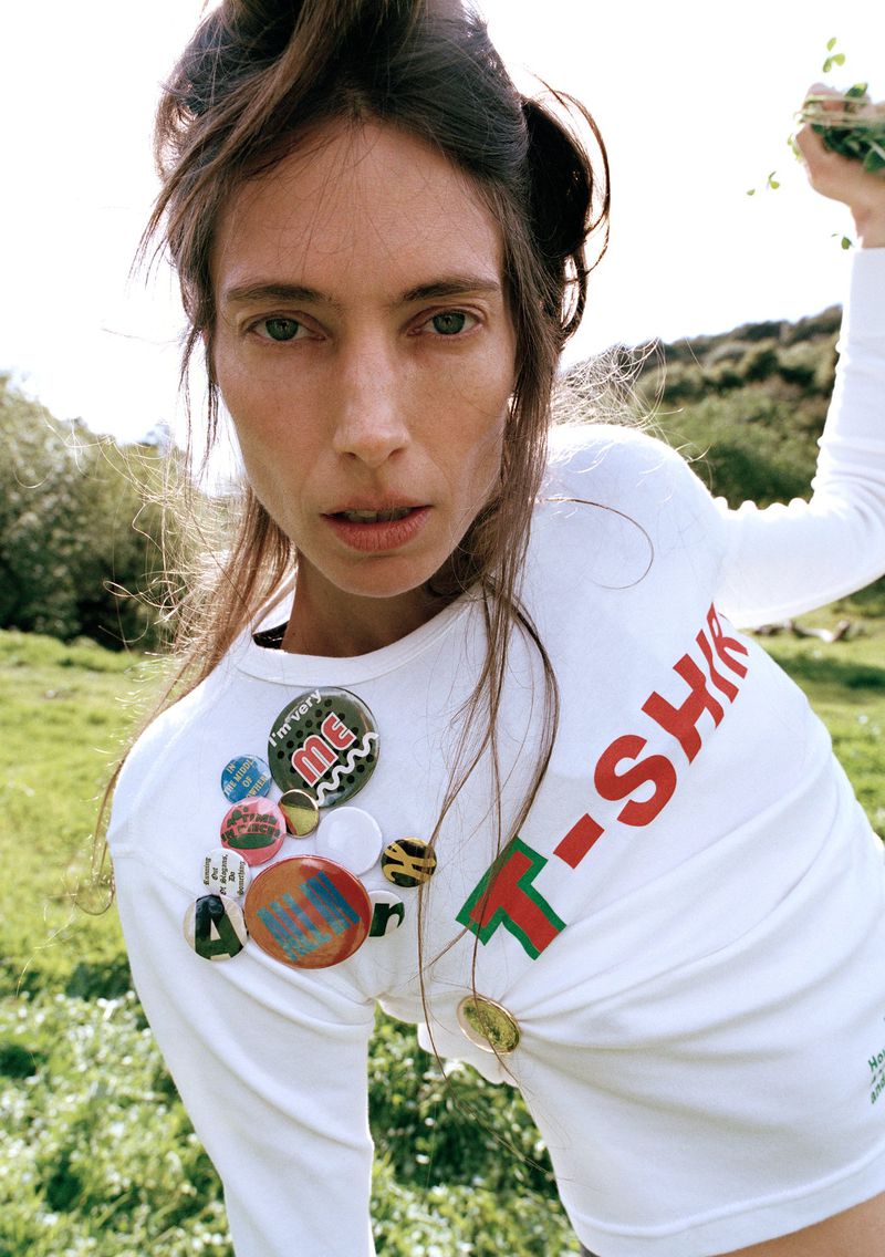 Woman in a field wearing a white long-sleeve T-shirt adorned with decorative pins and the word "T-SHIRT" in red and green lettering.