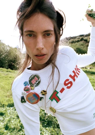 Woman in a field wearing a white long-sleeve T-shirt adorned with decorative pins and the word "T-SHIRT" in red and green lettering.
