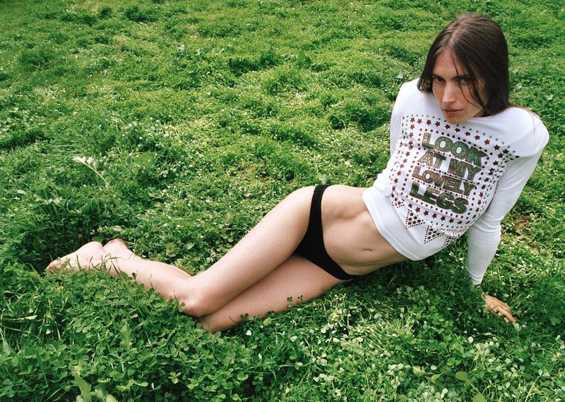 A young adult woman with long brown hair lying on a green grassy field. She is wearing a white long-sleeved shirt and black bikini bottoms.