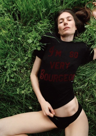 Model Jessica Miller wearing a black graphic t-shirt and briefs reclining on tall green grass outdoors.