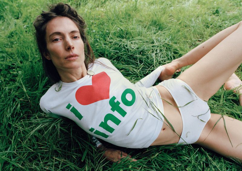 Woman in a white graphic top with red heart and green text poses in the grass outdoors in a medium shot with natural lighting.
