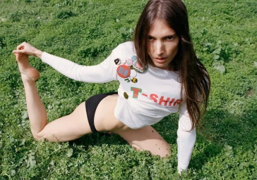Model Jessica Miller posing in a white long-sleeve crop top with badges and black briefs on a green grass field.