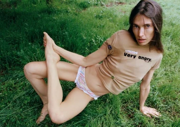 Jessica Miller wearing a tan graphic t-shirt and striped ruffled briefs reclining barefoot on green grass in natural light.