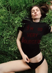 Model Jessica Miller wearing a black graphic t-shirt and briefs reclining on tall green grass outdoors.