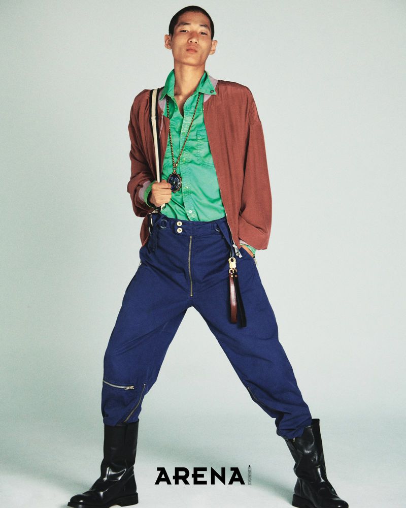 Model Bojvck Kang wearing a green shirt, rust brown jacket, cobalt blue trousers, and black boots against a pale seamless studio backdrop.