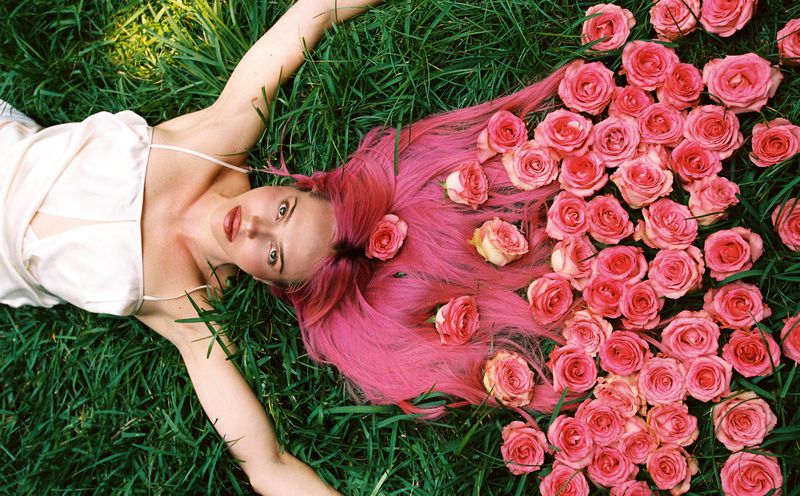Woman with long bright pink hair lying on green grass surrounded by scattered pink roses, wearing a white satin halter top.