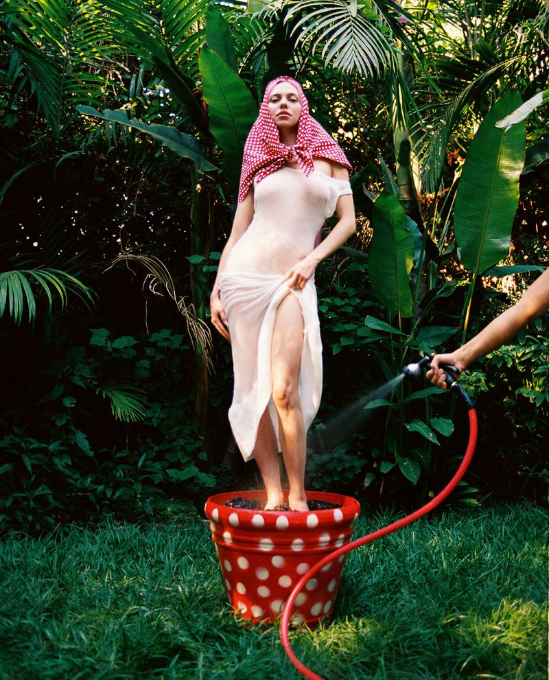 A woman wearing a sheer white dress and red headscarf stands inside a red polka-dot flowerpot in a garden while being sprayed with a hose.