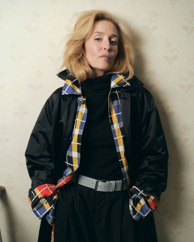 Gillian Anderson wearing a black coat with colourful plaid lining and a grey belt against floral wallpaper.