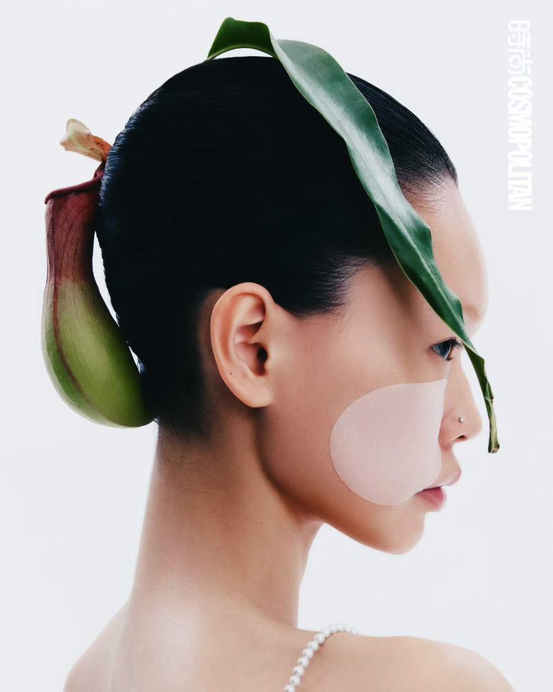 Profile portrait of model Saien Li with a green pitcher plant nestled in her hair and a pink cheek patch against a white background.