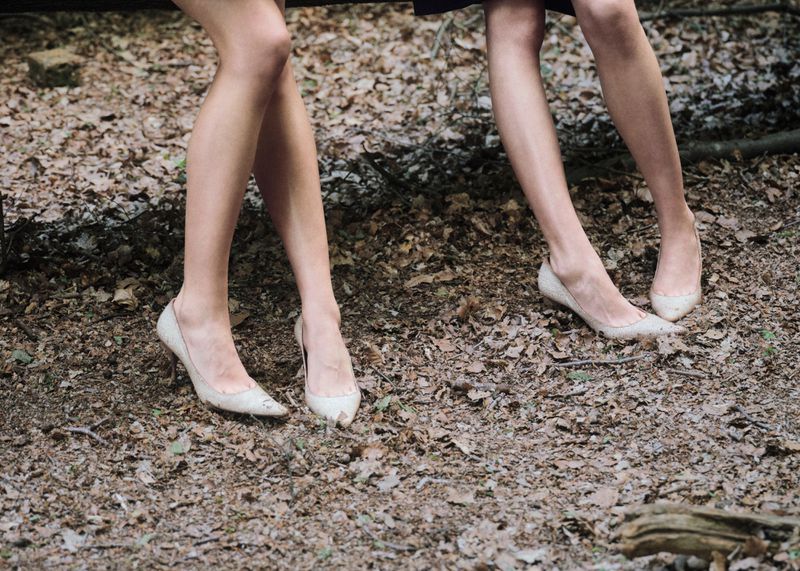 Two pairs of women's legs wearing off-white pointed-toe high heels on a forest floor covered in dry leaves.