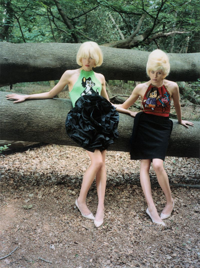 Two blonde models in graphic halter tops and black skirts pose leaning against a fallen log in a dense forest.