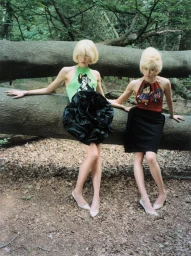 Two blonde models in graphic halter tops and black skirts pose leaning against a fallen log in a dense forest.
