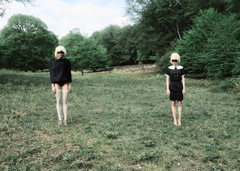 Two models with blonde wigs and eye coverings standing in a grassy forest clearing wearing black and white fashion garments.