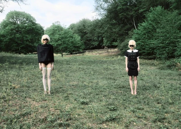Two models with blonde wigs and eye coverings standing in a grassy forest clearing wearing black and white fashion garments.