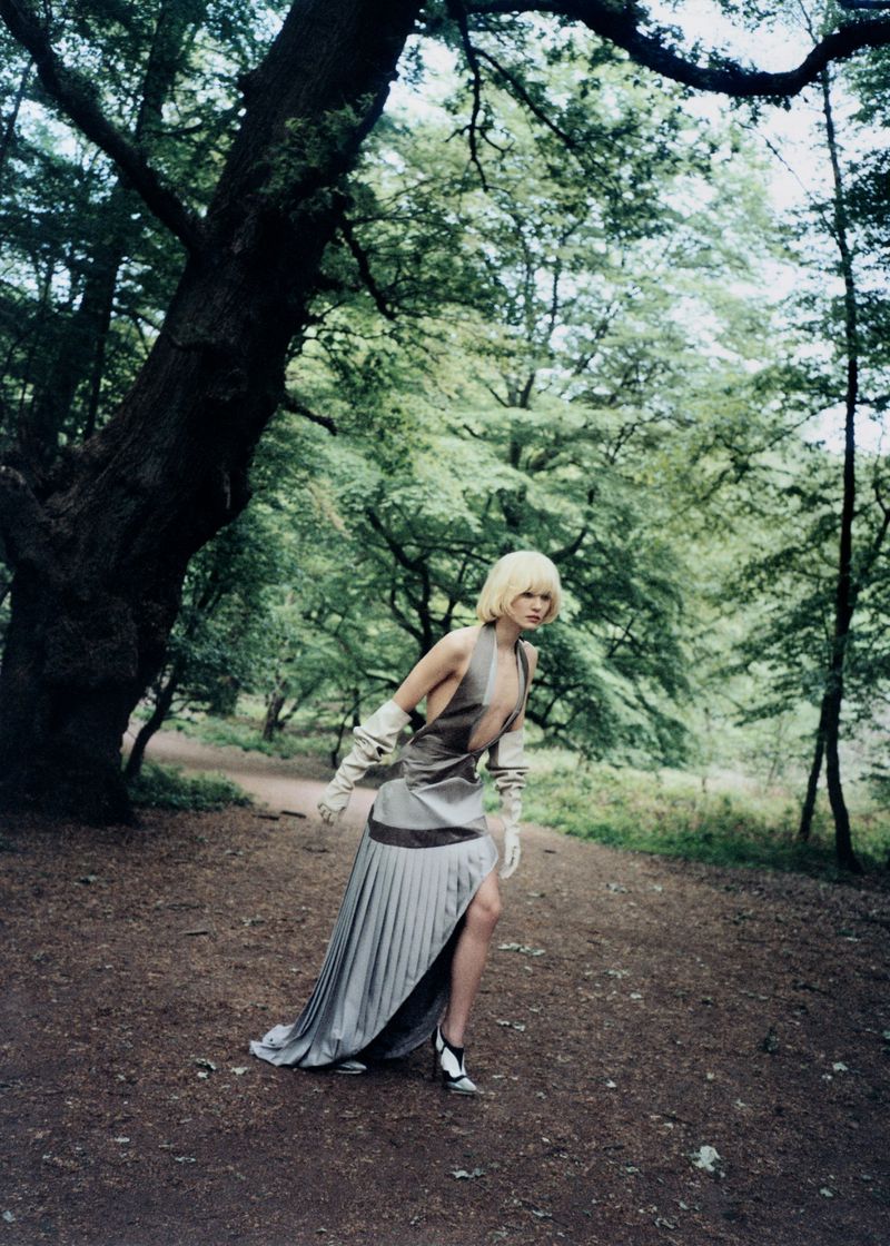 Model with a blonde bob posing in a pleated silver halter dress and long white gloves along a woodland trail.