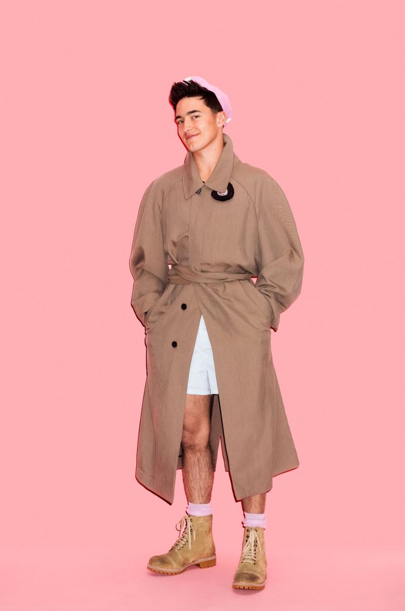 Man wearing a brown trench coat, light-colored shorts, and beige lace-up boots, posing against a solid pink backdrop.