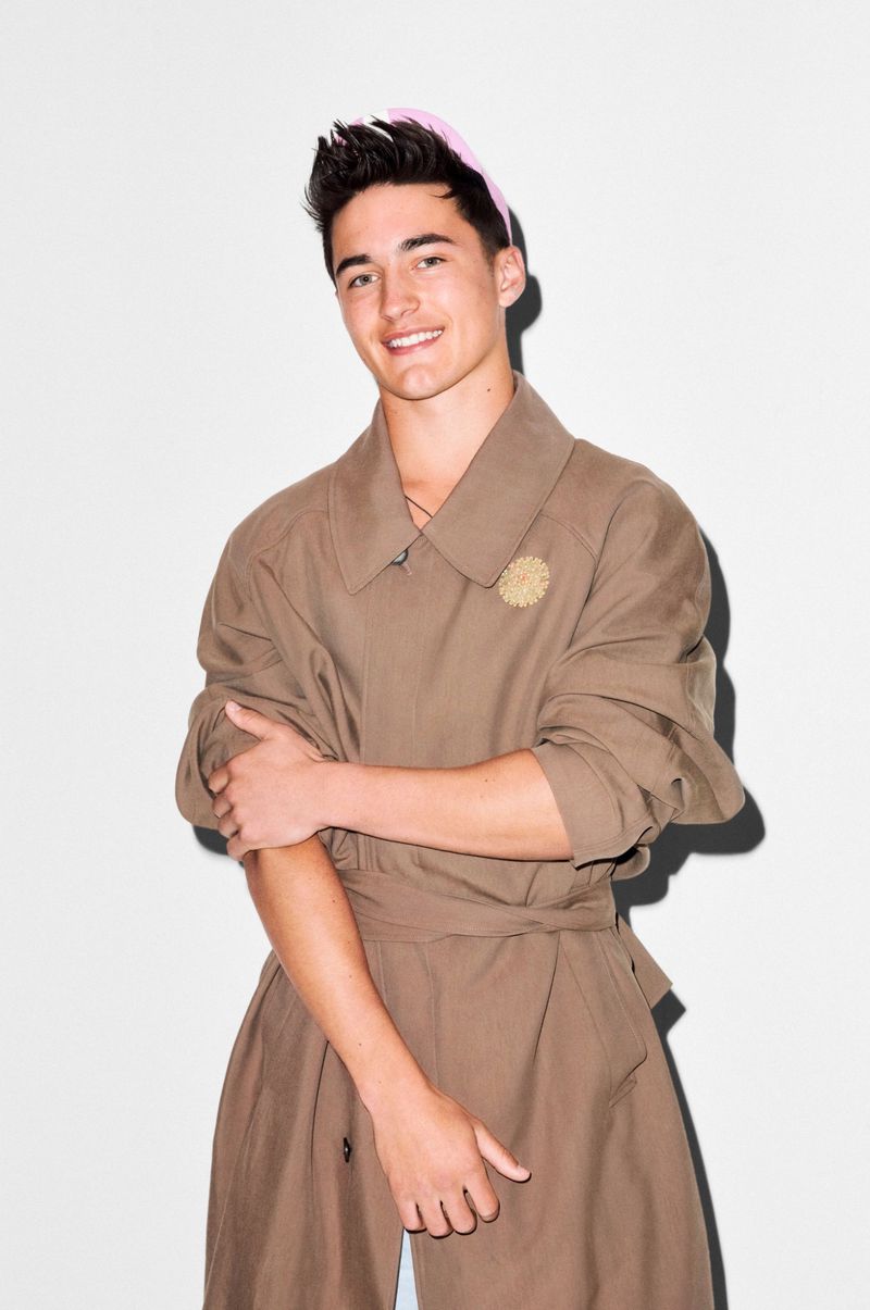 Model Lincoln Donald wearing an oversized belted taupe trench coat and a light pink cap against a white studio wall with hard shadow.