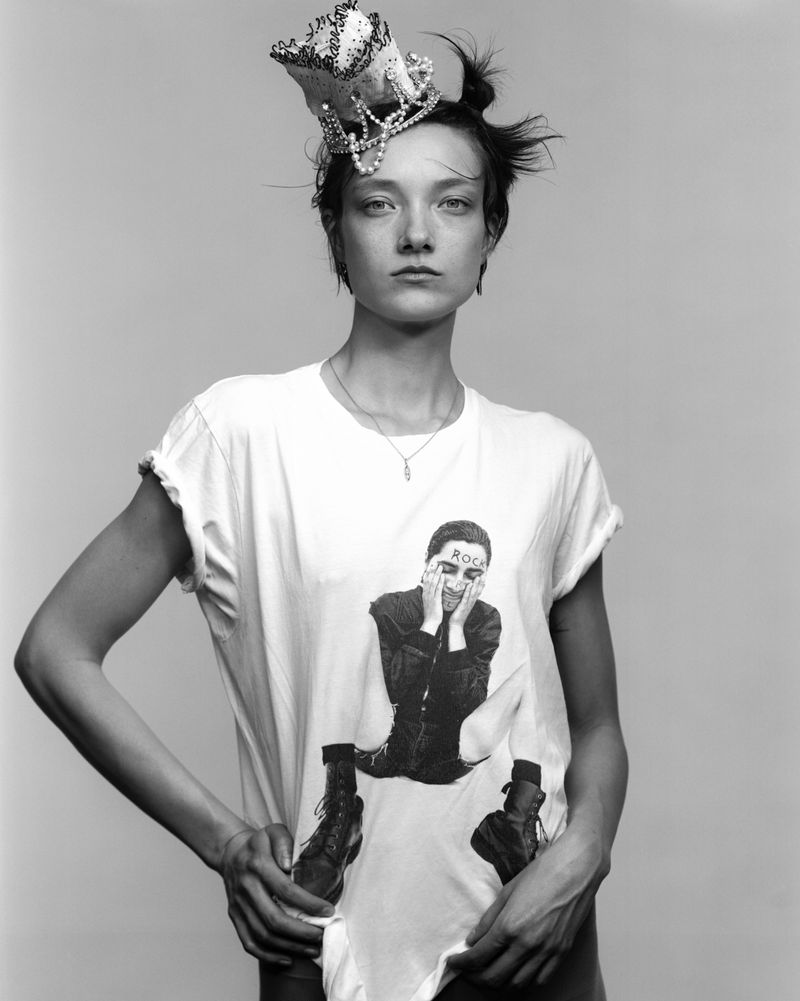 Model Yumi Lambert wearing a beaded crown headpiece and graphic white t-shirt against a grey studio backdrop.