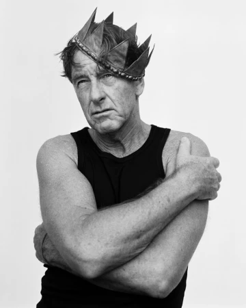 Man wearing a metal crown and black tank top with arms crossed against a plain white background