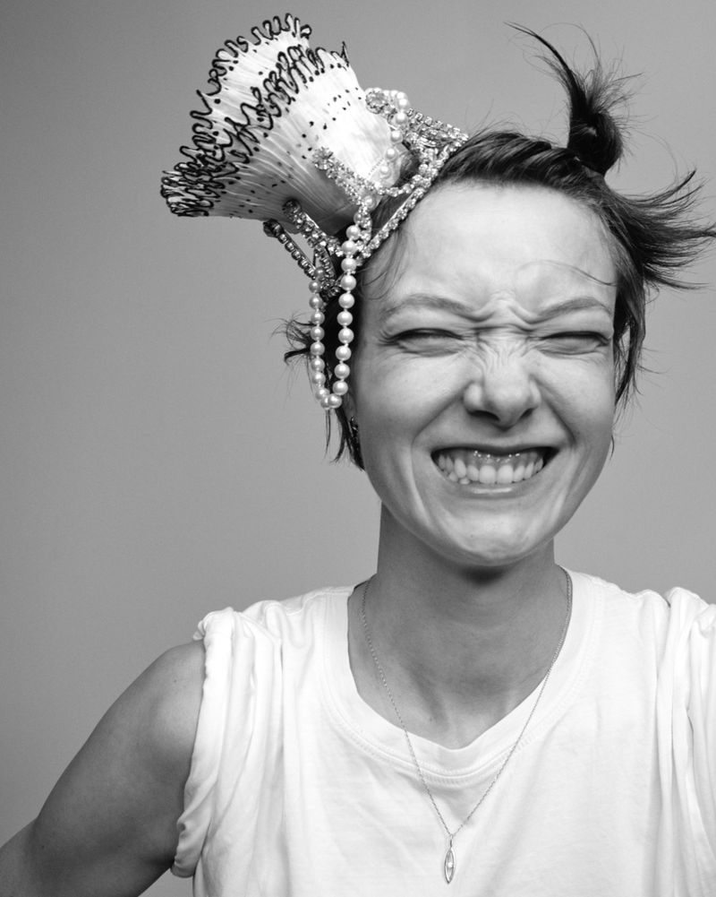 Black and white portrait of a woman laughing with eyes closed, wearing an ornate pearl crown headpiece and white sleeveless top.