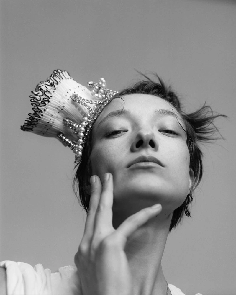 Low-angle black and white portrait of model Yumi Lambert wearing an ornate pearl and crystal headpiece while touching her cheek.