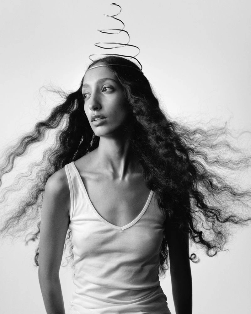 Monochrome portrait of a woman with long wavy windswept hair wearing a wire spiral crown and a white tank top.