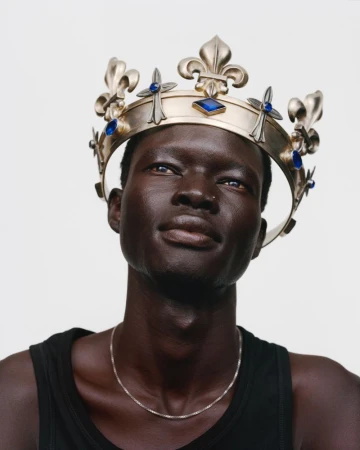 Close-up portrait of a Black man wearing an ornate gold fleur-de-lis crown with blue jewels and a black tank top against a white backdrop.